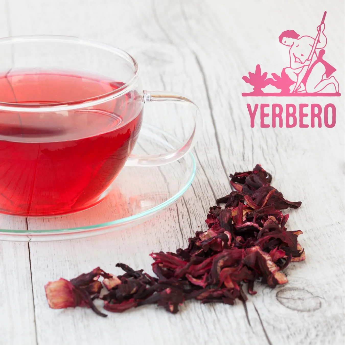 A clear glass cup of bright red hibiscus tea on a saucer next to a heap of dried hibiscus flowers