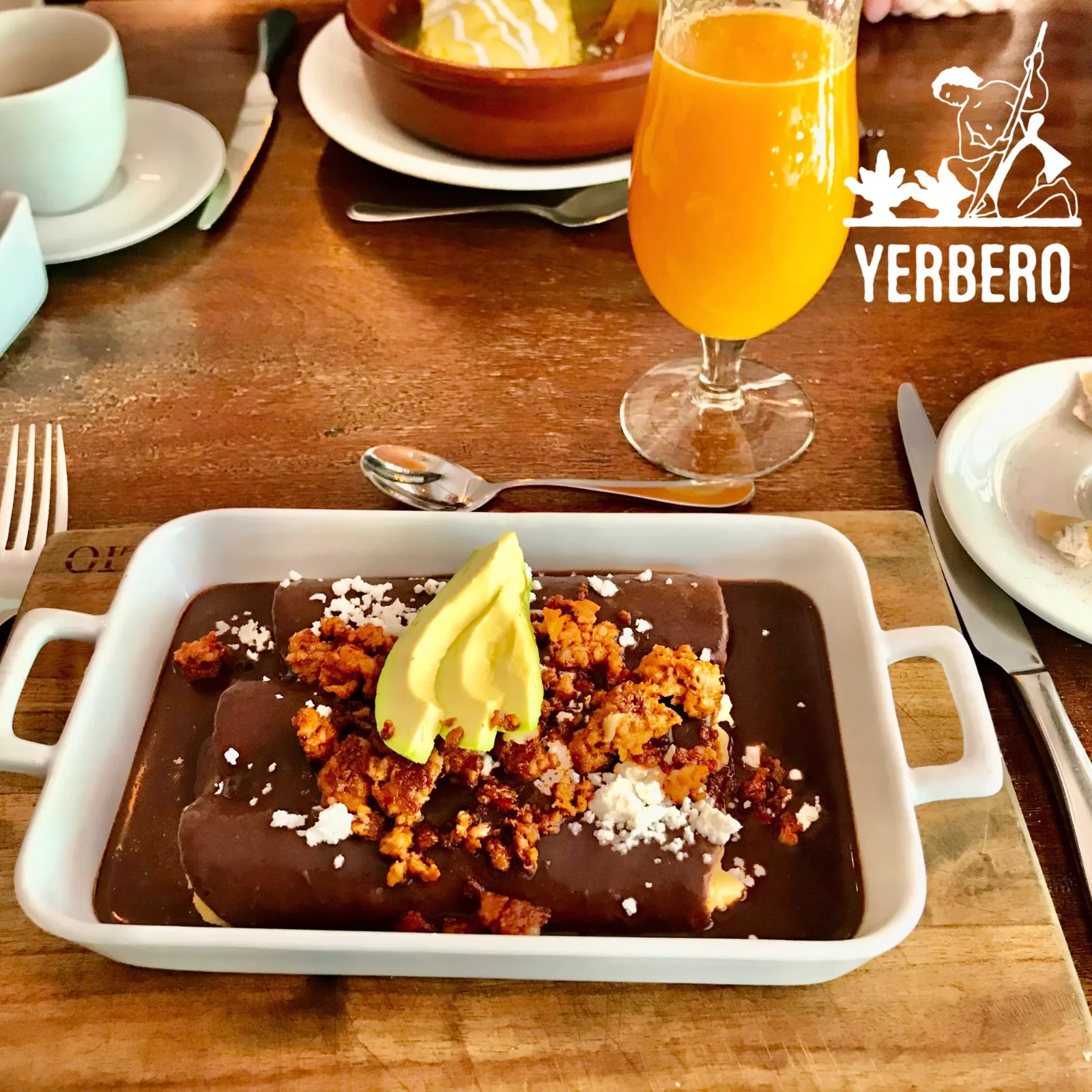 A white baking dish of tortillas covered in mole, topped with crumbled cheese and slices of avocado, beside a glass of orange juice